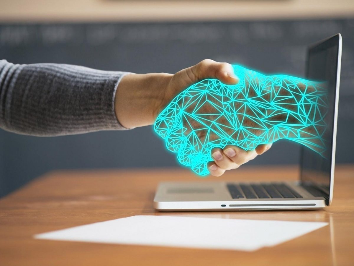 Poignée de main entre une main humaine et une main numérique sortant d'un ordinateur, symbole de la collaboration entre l'humain et l'intelligence artificielle
